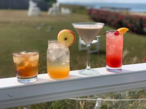 Four colorful cocktails on a railing, with a scenic outdoor background featuring grass and flowers.