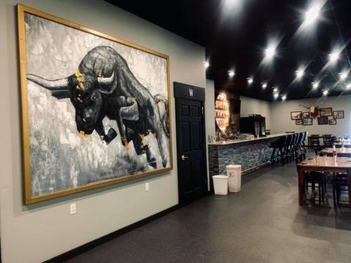 A large bull painting on a gray wall, with a bar and dining area visible in a modern restaurant setting.
