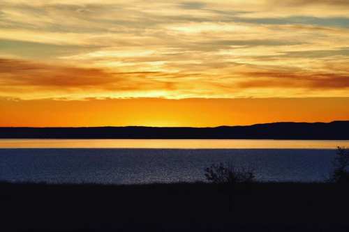 A serene sunset over a calm lake, with vibrant orange and yellow hues reflecting on the water and silhouetted hills in the background.