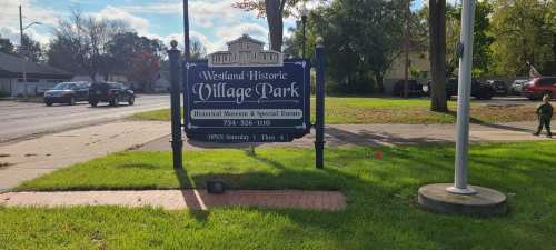 Sign for Westland Historic Village Park, with green grass and trees in the background, near a road.