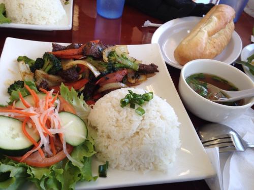 A plate of stir-fried vegetables and beef with rice, accompanied by a salad and a side of broth, with a bread roll.