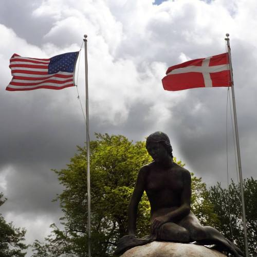 A statue of a seated figure with American and Danish flags waving above, set against a cloudy sky and green trees.