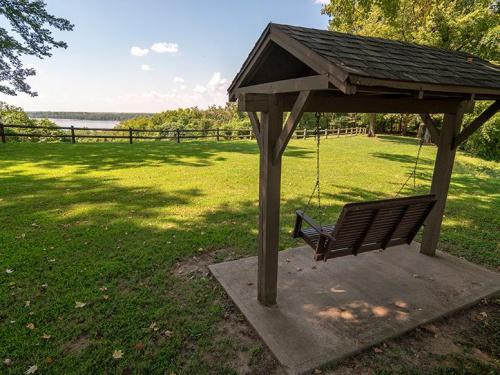 A wooden swing under a shelter overlooks a grassy area and a river in the distance, surrounded by trees.