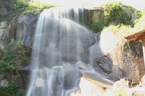 A cascading waterfall flows over rocky terrain, surrounded by lush greenery and sunlight.