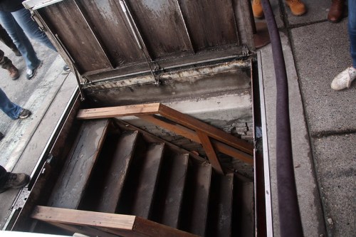 A view of wooden stairs leading down into a dark basement entrance, surrounded by concrete and wooden panels.