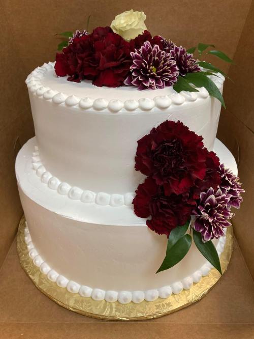 A two-tiered white cake adorned with red and purple flowers, presented in a cardboard box.