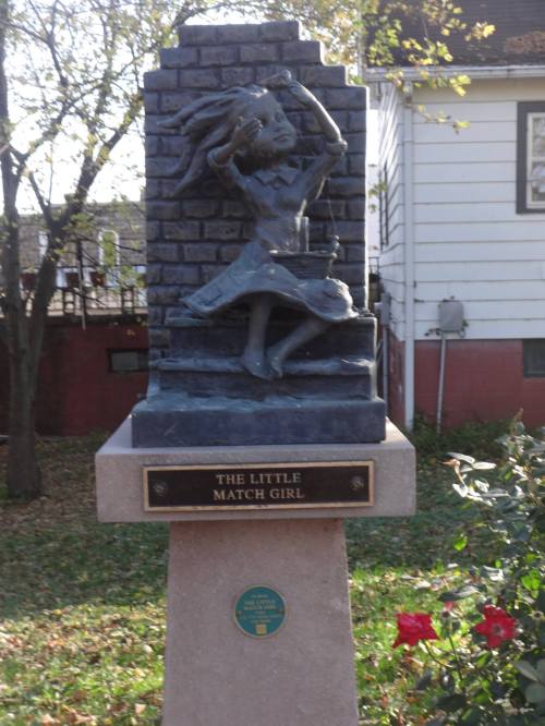 A bronze statue of a girl dancing, titled "The Little Match Girl," on a stone pedestal surrounded by greenery.