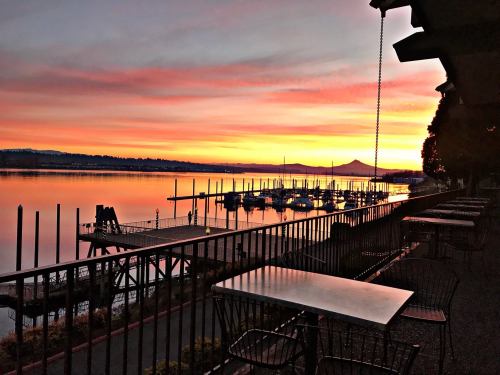 Sunset over a calm harbor with boats docked, reflecting vibrant colors in the water, and tables set along the shore.