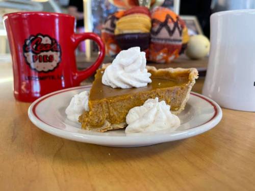 A slice of pumpkin pie topped with whipped cream on a plate, with coffee mugs in the background.