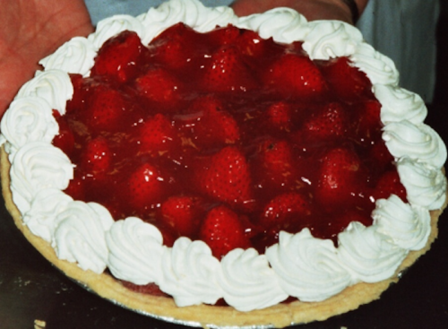 A strawberry pie topped with whipped cream and a glossy red glaze, held by a person.