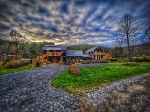 A scenic view of a modern house with wooden decks, surrounded by greenery and a gravel path under a cloudy sky.