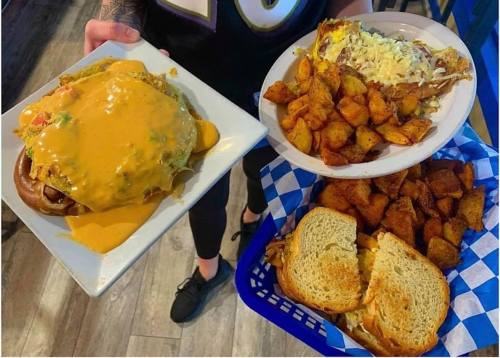 A person holds two plates: one with a cheesy burger and the other with a sandwich and seasoned potato wedges.