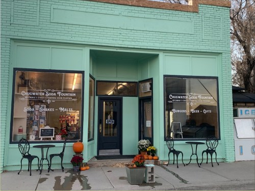 Exterior of Chugwater Soda Fountain, featuring large windows, a turquoise facade, and outdoor seating with pumpkins.