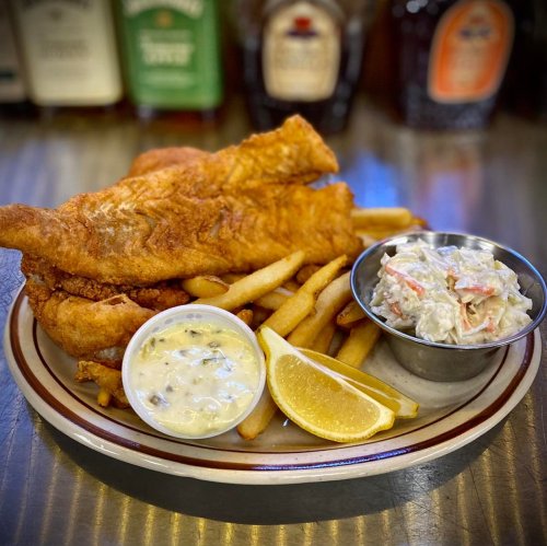 A plate of fried fish fillets, French fries, coleslaw, and a lemon wedge, with sauces on the side.