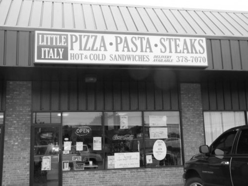 Black and white image of a restaurant storefront with a sign for "Little Italy" offering pizza, pasta, and steaks.