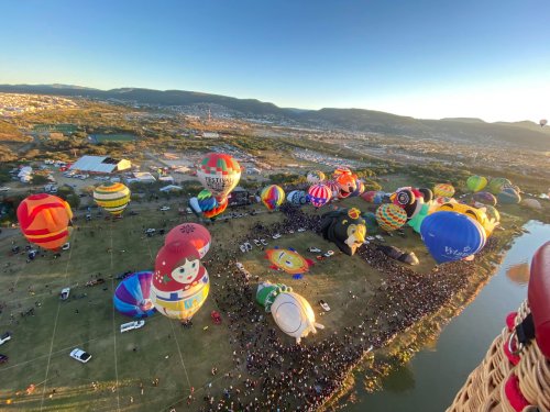 Aerial view of colorful hot air balloons over a crowd at a festival, with a scenic landscape in the background.