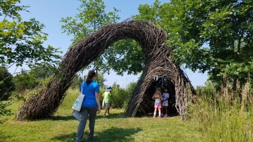 A large, arching structure made of branches, with people exploring inside and around it in a grassy area.