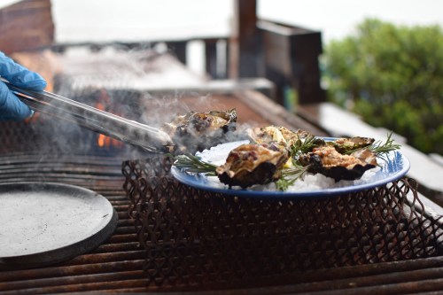 A hand with tongs lifts grilled oysters from a smoky grill onto a blue plate with herbs and ice.