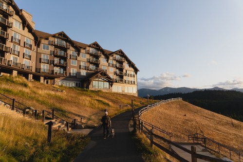 A person walks a dog along a path beside a large hotel, with rolling hills and mountains in the background at sunset.