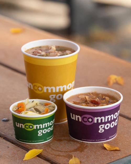 Three cups of soup in colorful containers sit on a wooden table, surrounded by autumn leaves.