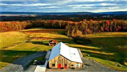 Aerial view of a rustic barn surrounded by colorful autumn foliage and rolling hills under a cloudy sky.