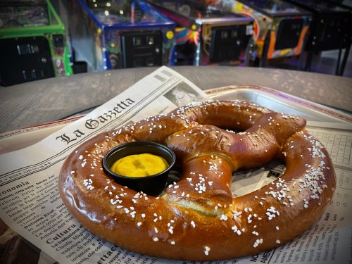 A large pretzel with salt served on a newspaper, accompanied by a small cup of mustard, with pinball machines in the background.
