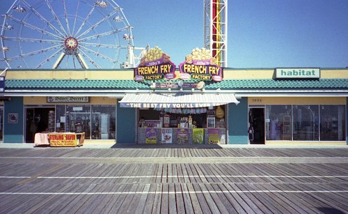 A colorful boardwalk eatery with a sign reading "French Fry Factory" and a ferris wheel in the background.