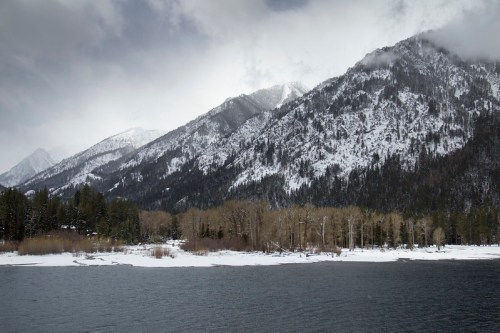 Snow-capped mountains rise above a serene lake, surrounded by trees and a cloudy sky.