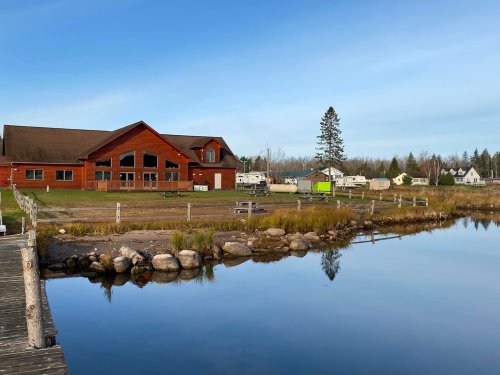 A serene lakeside view featuring a red building, grassy area, and reflections on calm water under a clear blue sky.
