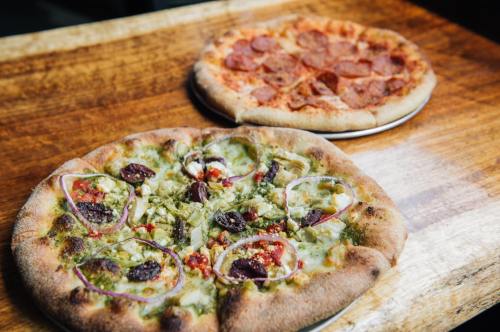 Two pizzas on wooden table: one with pepperoni and the other topped with green pesto, olives, and red onions.