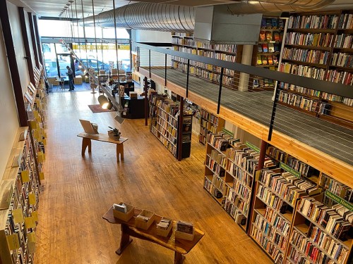 A spacious bookstore with wooden floors, tall bookshelves, and a second-level balcony overlooking the reading area.