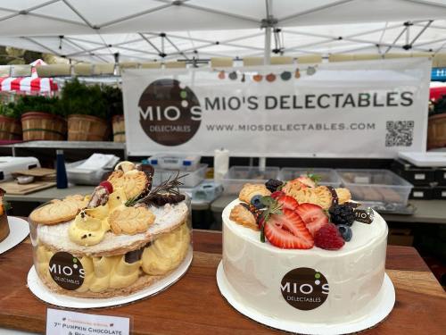 Two beautifully decorated cakes on display at a market stall, with a sign reading "Mio's Delectables" in the background.