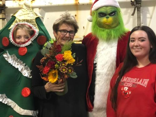 A group of four people in festive costumes, including a Christmas tree and the Grinch, holding flowers and smiling.
