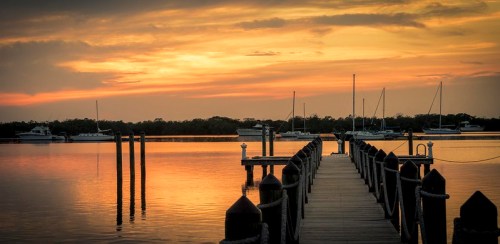 A serene sunset over a calm waterway, with boats docked and a wooden pier extending into the scene.