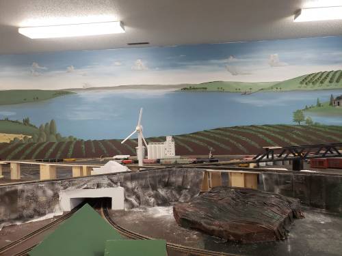A model train setup featuring a painted landscape with a lake, vineyards, and a wind turbine in the background.