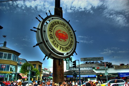 Sign for Fisherman's Wharf in San Francisco, surrounded by a bustling crowd and shops under a blue sky.