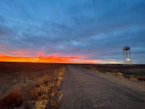 A dirt road leads to a water tower against a vibrant sunset with colorful clouds in the sky.