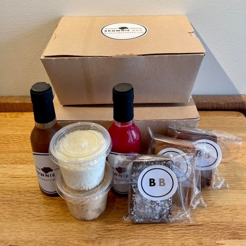 Two boxes from Brownie Bar with sauces, dessert cups, and individually wrapped brownies on a wooden surface.