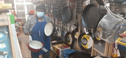 A person wearing a mask and gloves holds a pot in a kitchenware store, surrounded by various cooking pans.