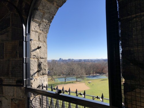 View from a stone archway overlooking a park with trees and a river under a clear blue sky.
