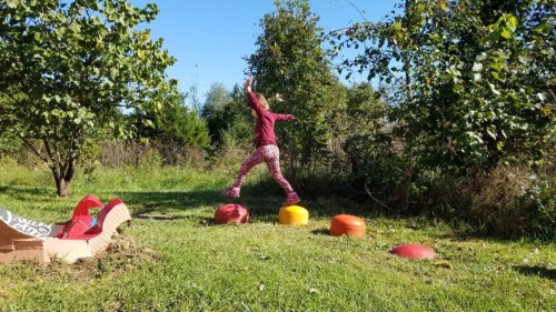 A child in colorful clothing jumps over colorful stepping stones in a grassy area surrounded by trees.