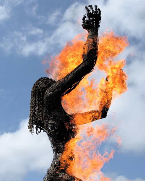 A towering metal sculpture of a woman with flames erupting from her hands against a cloudy sky.
