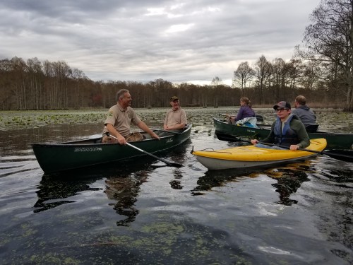 A group of people kayaking and canoeing on a calm lake surrounded by trees and cloudy skies.