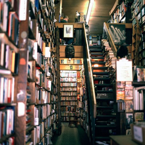 A cozy bookstore with tall shelves filled with books, a staircase leading up, and warm lighting creating an inviting atmosphere.