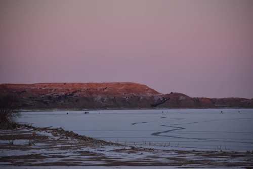 A serene winter landscape featuring a frozen lake and distant hills at dusk, with soft pink and purple hues in the sky.