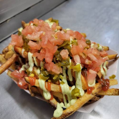 A serving of fries topped with diced tomatoes, pickles, and drizzled with sauce on a metal surface.