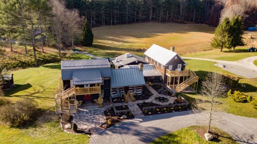 Aerial view of a modern house with multiple sections, surrounded by greenery and a gravel driveway.