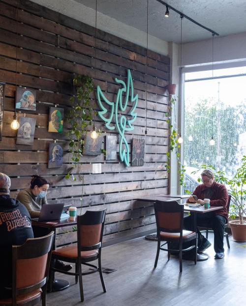 A cozy café interior with wooden walls, neon art, and patrons enjoying coffee at tables surrounded by plants.