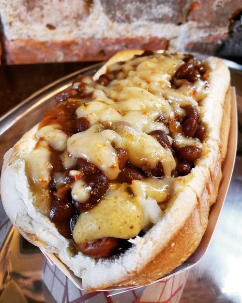 A loaded hot dog topped with melted cheese, chili, and beans, served in a soft bun on a metal tray.