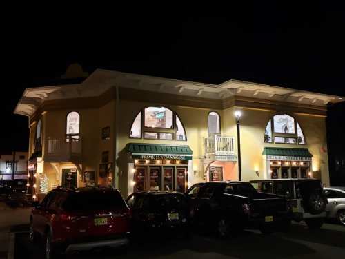 A brightly lit restaurant building at night, featuring large windows and outdoor seating, surrounded by parked cars.